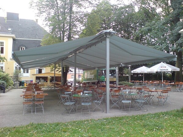 Überdachter Biergarten mit vielen leeren Holztischen und Stühlen im Grünen.