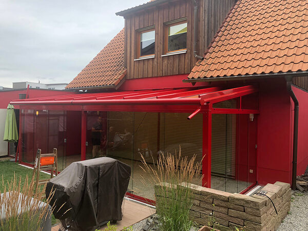 Modernes Einfamilienhaus mit roter Glas-Terrassenüberdachung und Gartenmöbeln.