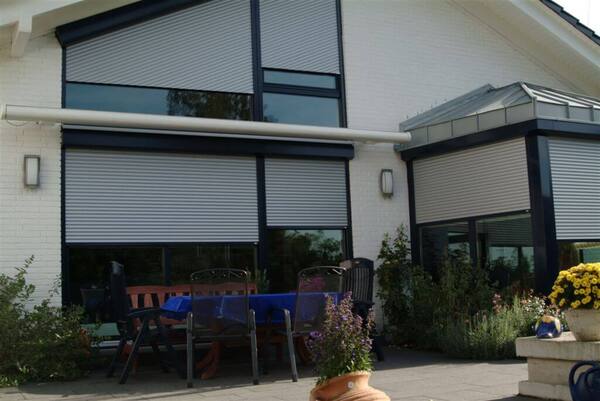 Moderne Terrasse mit Gartenmöbeln vor Haus mit geschlossenen Rollläden und Blumen.