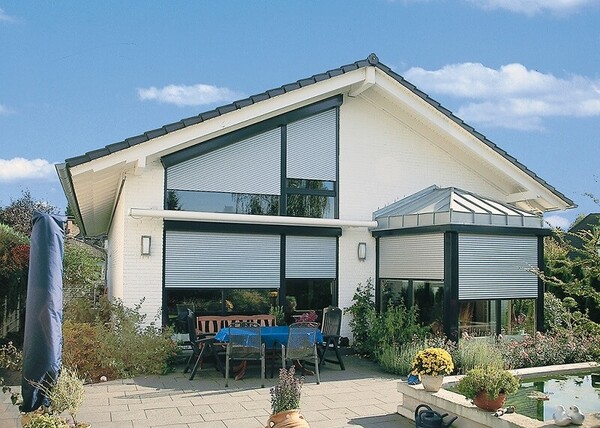 Modernes Einfamilienhaus mit Rollläden, Terrasse und Garten bei sonnigem Wetter.