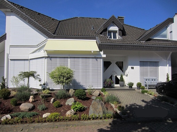 Modernes Einfamilienhaus mit gepflegtem Vorgarten, Markise und Jalousien bei Sonnenschein.