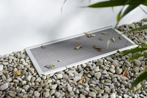 Lichtschachtabdeckung aus Metall mit Laubschutzgitter auf Kiesboden an Hauswand.