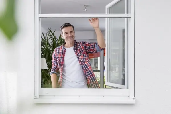 Mann lüftet frische Luft durch geöffnetes Fenster in modernem, hellen Wohnzimmer.